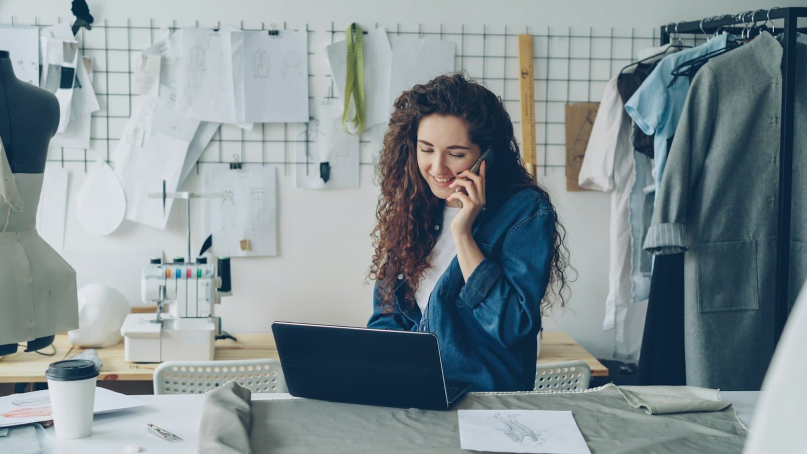 A small business owner on the phone at her workbench, laptop open, orders piling up around her