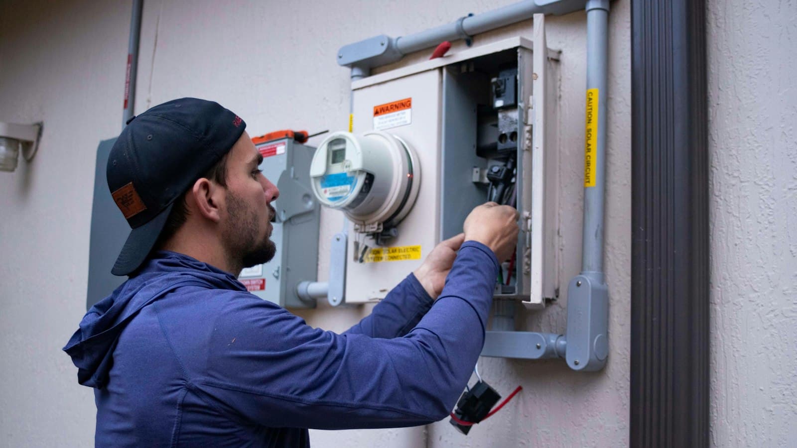 A technician in a cap working on a meter box outside a building — the owner in the field is not answering the phone